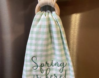 May include: A green and white checkered kitchen towel with a fringe border hangs from a wooden ring. The towel has the text "Spring is here!" embroidered in green thread.