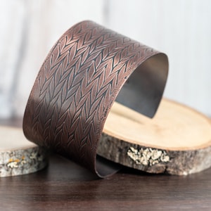 May include: A copper cuff bracelet with a woven leaf pattern. The bracelet is on a wooden surface.