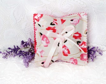 May include: A small, square-shaped pillow with a floral print in shades of pink, white, and green. The pillow is tied with a pink ribbon and has a scalloped edge.