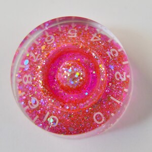 Peut inclure: Un dé rond et transparent avec un motif en spirale rose et orange pailleté. Des chiffres blancs sont visibles sur le bord. Accessoire de jeu.