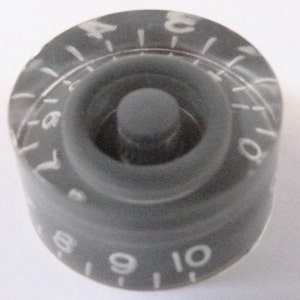 Pode incluir: Botão de guitarra cinza escuro com marcações numéricas brancas de 0 a 10. O botão tem um design circular com um centro elevado e uma borda externa canelada. Os números são espaçados uniformemente.