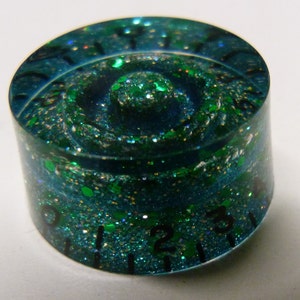 Pode incluir: Uma maçaneta azul-esverdeada e verde com glitter e números pretos 1, 2 e 3 ao redor da borda.