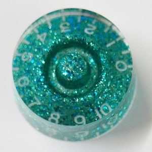 May include: A teal blue round knob with white numbers 0-12 around the edge. The knob is covered in glitter and has a smaller circle in the center with a teal blue glitter centre.