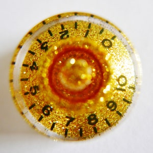 Pode incluir: Uma maçaneta dourada com números pretos de 4 a 10 ao redor da borda. O centro da maçaneta é um design de glitter dourado.