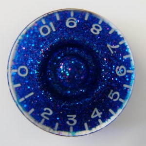 Peut inclure: Un bouton bleu avec des chiffres blancs de 1 à 10 et de 4 à 8. Le bouton est recouvert de paillettes bleues avec des paillettes roses et violettes.
