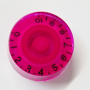 May include: A vibrant pink guitar volume knob with black numerals from 0 to 10. The circular knob features a central recess and a ridged edge. The numbers are evenly spaced around the circumference.