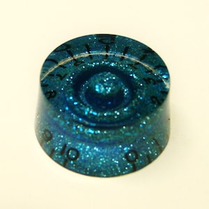 Pode incluir: Uma botão de guitarra azul turquesa com acabamento brilhante e números pretos de 0 a 10.