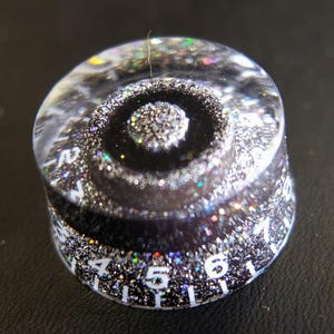 Pode incluir: Um botão de guitarra transparente preenchido com glitter, com um centro preto e números brancos. O botão redondo tem uma superfície texturizada e um efeito cintilante e iridescente. Os números de 1 a 7 são visíveis.