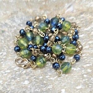 May include: A gold-toned necklace with a lobster clasp. The necklace features a mix of green and blue marbled beads, small dark blue beads, and gold-colored beads. The beads are strung on a delicate gold chain.