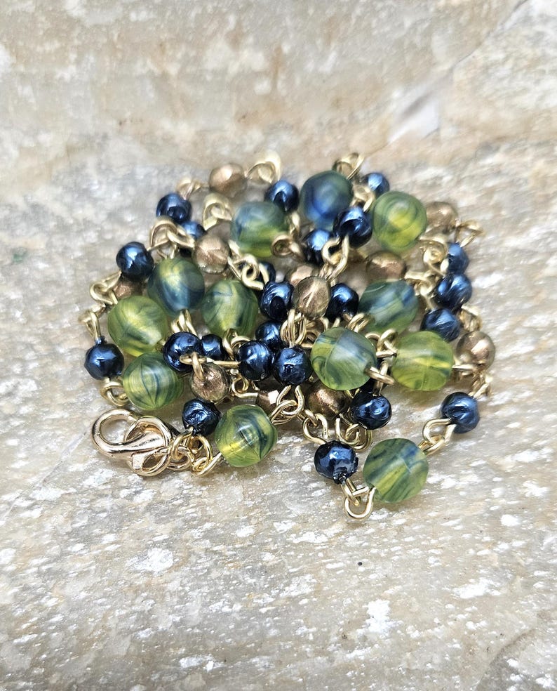 May include: A gold-toned necklace with a lobster clasp. The necklace features a mix of green and blue marbled beads, small dark blue beads, and gold-colored beads. The beads are strung on a delicate gold chain.