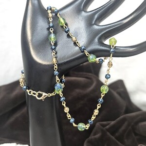 May include: A delicate necklace featuring a gold chain with alternating beads in shades of green, blue, and brown. The necklace is displayed on a black hand-shaped jewelry holder, showcasing its intricate design and color palette. The clasp is gold.