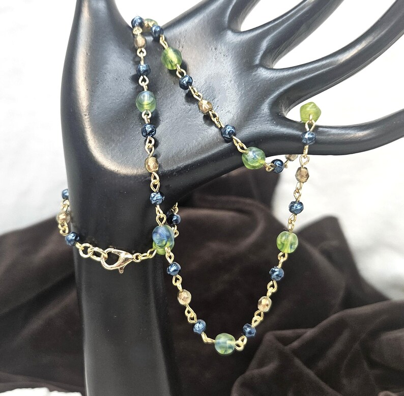 May include: A delicate necklace featuring a gold chain with alternating beads in shades of green, blue, and brown. The necklace is displayed on a black hand-shaped jewelry holder, showcasing its intricate design and color palette. The clasp is gold.