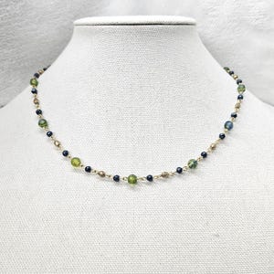 May include: Delicate gold-tone necklace featuring a strand of small, dark blue, olive green, and gold-colored beads. The necklace is displayed on a white mannequin bust, showcasing its design and color variations.