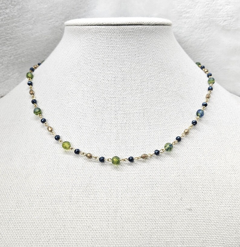 May include: Delicate gold-tone necklace featuring a strand of small, dark blue, olive green, and gold-colored beads. The necklace is displayed on a white mannequin bust, showcasing its design and color variations.
