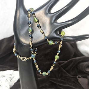 May include: A delicate necklace with a gold chain and a series of small, colorful beads. The beads are a mix of green, blue, and tan, creating a subtle yet eye-catching design. The necklace is displayed on a black hand-shaped jewelry holder.