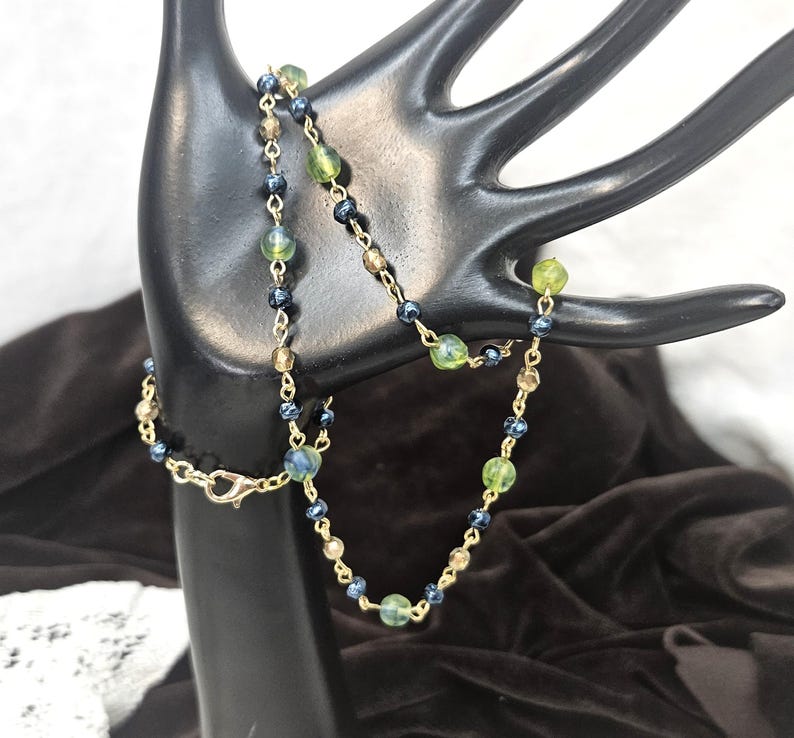 May include: A delicate necklace with a gold chain and a series of small, colorful beads. The beads are a mix of green, blue, and tan, creating a subtle yet eye-catching design. The necklace is displayed on a black hand-shaped jewelry holder.