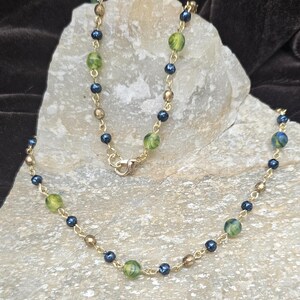 May include: A gold-colored necklace with a delicate chain. The necklace features a pattern of small, dark blue beads, interspersed with larger, green and gold-colored beads. The necklace is displayed on a light-colored stone.