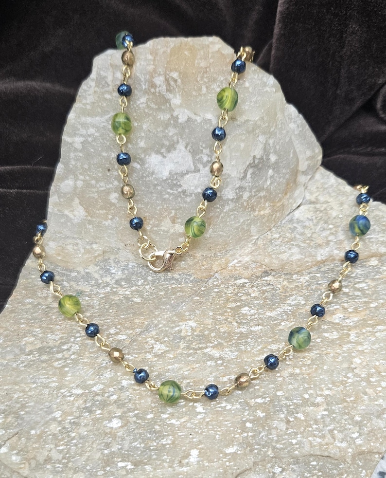 May include: A gold-colored necklace with a delicate chain. The necklace features a pattern of small, dark blue beads, interspersed with larger, green and gold-colored beads. The necklace is displayed on a light-colored stone.