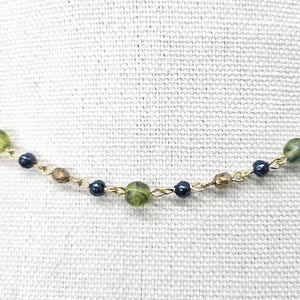 May include: A delicate necklace featuring a gold chain with alternating beads. The beads include green, dark blue, and brown tones. The necklace is displayed against a textured, off-white background.