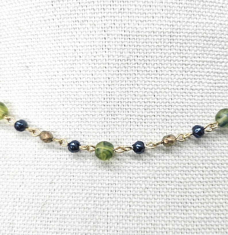 May include: A delicate necklace featuring a gold chain with alternating beads. The beads include green, dark blue, and brown tones. The necklace is displayed against a textured, off-white background.