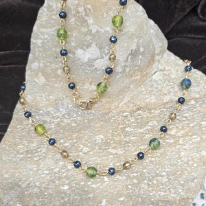 May include: A gold-toned necklace with a delicate chain. The necklace features a pattern of small, dark blue beads, interspersed with larger, green and gold-colored beads. The necklace is displayed on a light-colored stone.