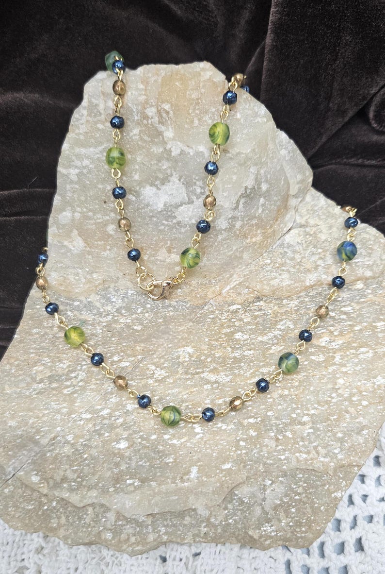 May include: A gold-toned necklace with a delicate chain. The necklace features a pattern of small, dark blue beads, interspersed with larger, green and gold-colored beads. The necklace is displayed on a light-colored stone.