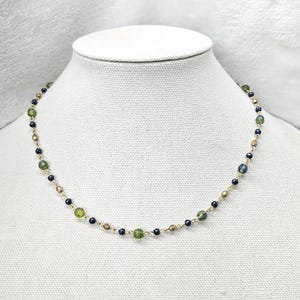 May include: A delicate necklace featuring a gold chain with alternating green, dark blue, and gold-colored beads. The necklace is displayed on a white mannequin bust, showcasing its design and color palette.