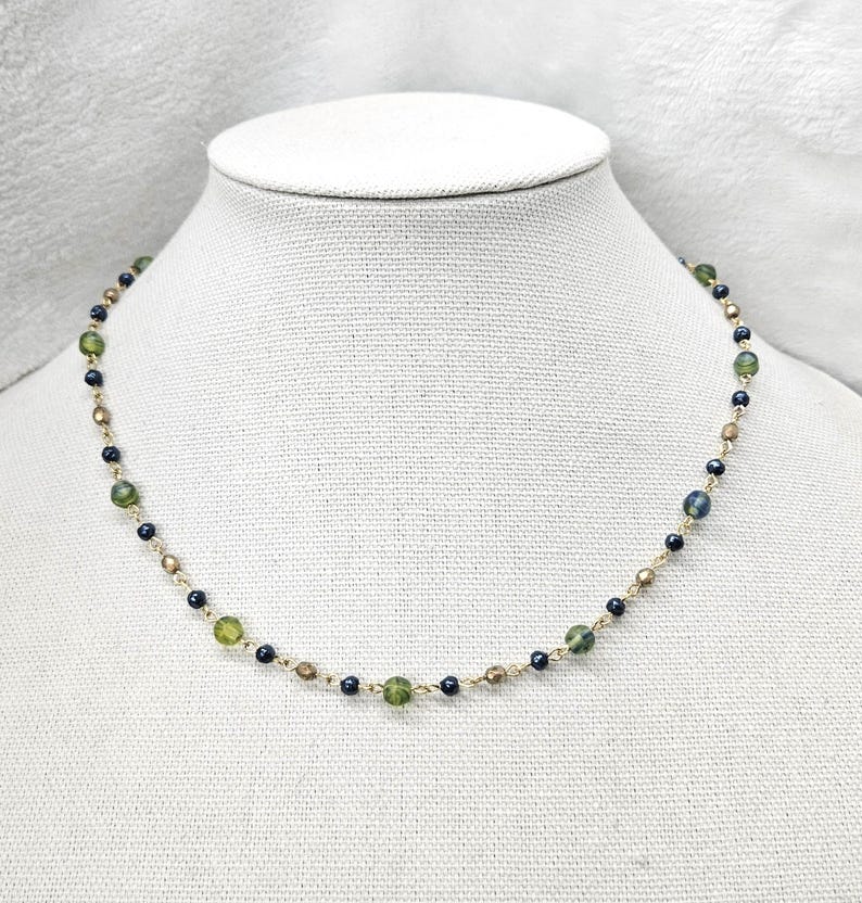 May include: A delicate necklace featuring a gold chain with alternating green, dark blue, and gold-colored beads. The necklace is displayed on a white mannequin bust, showcasing its design and color palette.
