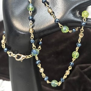 May include: A gold-colored chain necklace with a lobster clasp, adorned with a mix of green, blue, and gold-toned beads. The beads vary in size and shape, creating a delicate and elegant design. The necklace is displayed against a dark background.