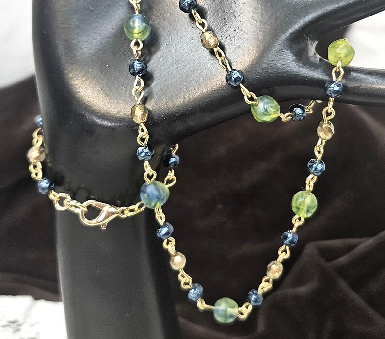 May include: A gold-colored chain necklace with a lobster clasp, adorned with a mix of green, blue, and gold-toned beads. The beads vary in size and shape, creating a delicate and elegant design. The necklace is displayed against a dark background.
