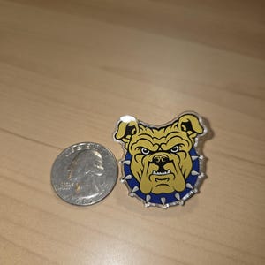 May include: A yellow and blue bulldog pin with a silver quarter for size comparison. The pin features a bulldog head with a spiked collar. The bulldog has a stern expression.
