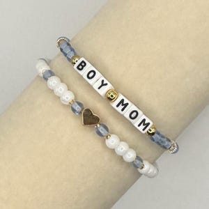May include: A set of two beaded bracelets. One bracelet features white square beads with black letters spelling BOY MOM, accented by gold-tone beads and blue-grey beads. The second bracelet features white round beads, blue-grey beads, and a gold-tone heart charm.