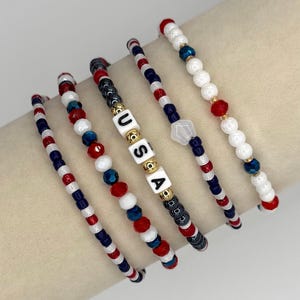 May include: A collection of beaded bracelets in red, white, and blue. One bracelet spells out "USA" with white letter beads. The bracelets feature a variety of bead sizes and shapes, including round and faceted beads. The bracelets are designed with patriotic colors.