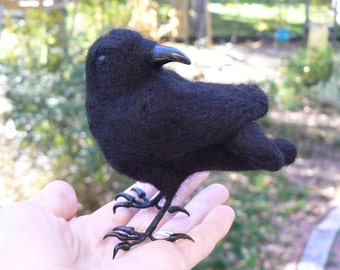 May include: A black felt crow with realistic details, including black eyes and a black beak. The crow is perched on a hand, with its feet visible.