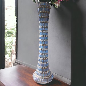 May include: A tall, blue and gold mosaic glass vase with a flared base. The vase is filled with pink flowers.