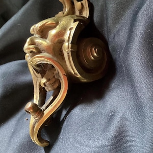 May include: Antique brass furniture hardware with a detailed face design. The hardware features a stylized face with intricate details, a scrolled design, and a circular element. The brass has a warm, aged patina.