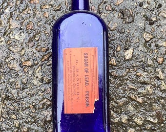 May include: A vintage, cobalt blue glass bottle with a rectangular shape and a dark blue stopper. A red label with black text reads "SUGAR OF LEAD - POISON" and details about the chemist. The bottle is likely from the late 19th or early 20th century.