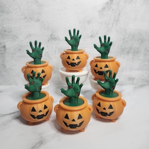 Könnte beinhalten: Orangefarbene Halloween-Dekotöpfe mit geschnitzten Kürbisgesichtern. Aus jedem Topf ragt eine grüne Hand heraus. Die Töpfe sind auf einer weißen Oberfläche angeordnet.