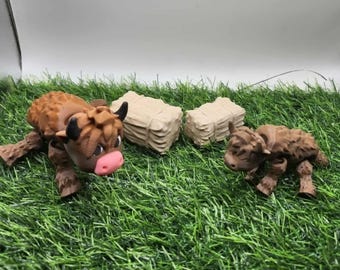 May include: A brown and tan Highland cow figurine with a pink nose and black horns, alongside a smaller calf figurine. Two hay bale figurines are also present, all set on a bed of green artificial grass. The figurines are likely 3D printed.