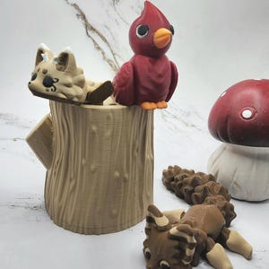 Puede incluir: Una escena caprichosa con una figura de cardenal rojo con pico naranja, una figura de zorro marrón y una figura de oruga marrón. El cardenal está posado sobre un tocón de árbol marrón, con una seta roja y blanca al fondo.