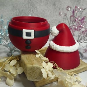 May include: A red Christmas-themed container and hat set. The container is designed to resemble Santa's belly, with a black belt and white buckle. The hat is red with a white trim and pom-pom. The set is placed on a wooden surface.
