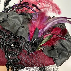 May include: A burgundy felt hat with black lace trim and a black ribbon. The hat is adorned with a cluster of feathers in shades of pink, purple, and black.