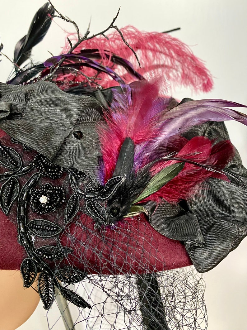 May include: A burgundy felt hat with black lace trim and a black ribbon. The hat is adorned with a cluster of feathers in shades of pink, purple, and black.