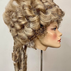 K&ouml;nnte beinhalten: Eine blonde Per&uuml;cke mit gro&szlig;en Locken, die zu einem hohen Dutt mit einem langen, gelockten Pferdeschwanz frisiert ist. Die Per&uuml;cke sitzt auf einem Mannequin-Kopf.