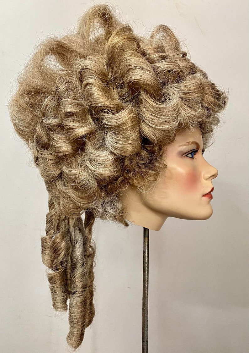 K&ouml;nnte beinhalten: Eine blonde Per&uuml;cke mit gro&szlig;en Locken, die zu einem hohen Dutt mit einem langen, gelockten Pferdeschwanz frisiert ist. Die Per&uuml;cke sitzt auf einem Mannequin-Kopf.