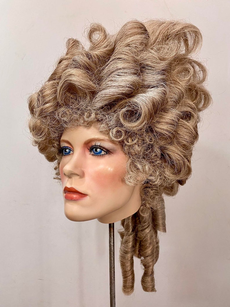 K&ouml;nnte beinhalten: Eine blonde Per&uuml;cke mit gro&szlig;en Locken und einem langen, gelockten Pferdeschwanz. Die Per&uuml;cke ist in einem klassischen, eleganten Stil gestylt.
