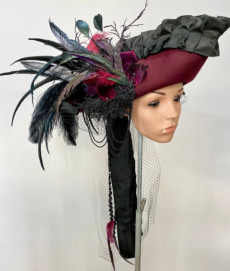 May include: A burgundy felt hat with a black feather plume and black velvet trim. The hat is adorned with black lace and beads, and a black veil hangs from the back.
