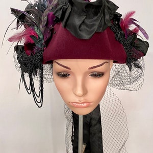 May include: A burgundy felt hat with a black ribbon and a black veil. The hat is decorated with black feathers, black lace, and a black bow. The hat is sitting on a mannequin head.
