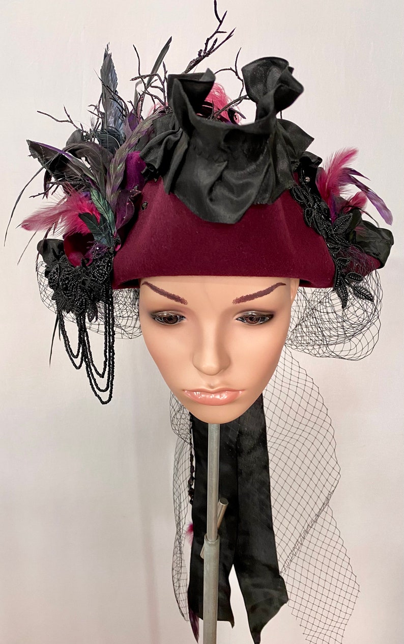 May include: A burgundy felt hat with a black ribbon and a black veil. The hat is decorated with black feathers, black lace, and a black bow. The hat is sitting on a mannequin head.