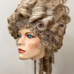 K&ouml;nnte beinhalten: Eine blonde Per&uuml;cke, die zu einer gro&szlig;en, aufwendigen Hochsteckfrisur mit Locken und Ringellocken gestylt ist. Die Per&uuml;cke wird auf einem Mannequin-Kopf pr&auml;sentiert.